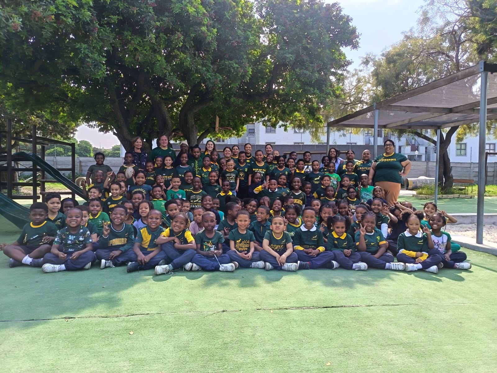 A large group of children and several adults pose for a group photo outdoors on a green surface, with trees and a building in the background. Most are wearing matching green and yellow shirts and smiling at the camera.