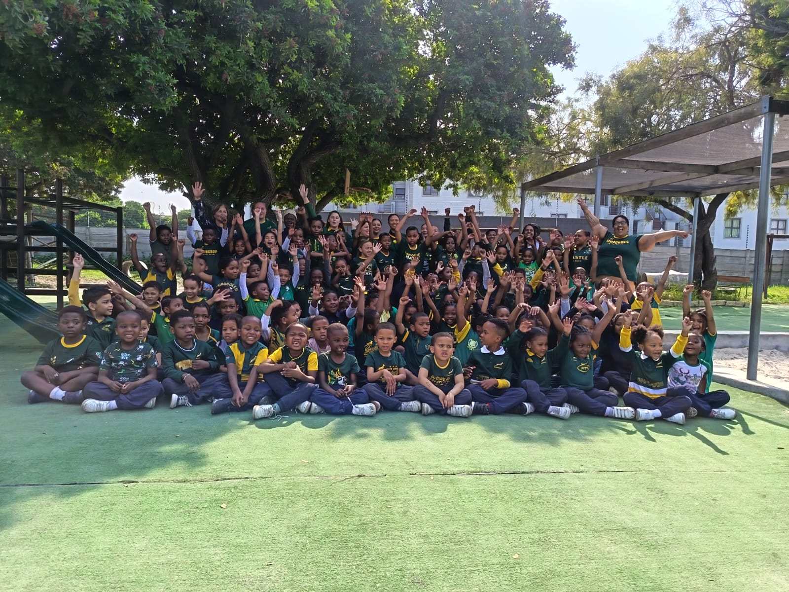 A large group of children and a few adults sit and stand outside on artificial grass, smiling and raising their arms in celebration. They are wearing coordinated dark green and yellow outfits. Trees and playground equipment are visible.