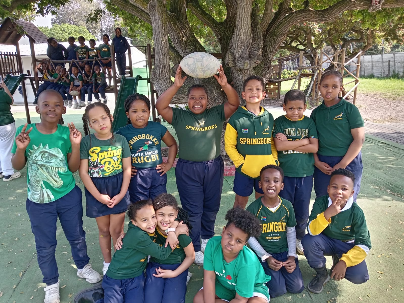 A group of schoolchildren in green and yellow rugby shirts pose outdoors under a tree, with one child holding a rugby ball above their head. More children are sitting on play equipment in the background.