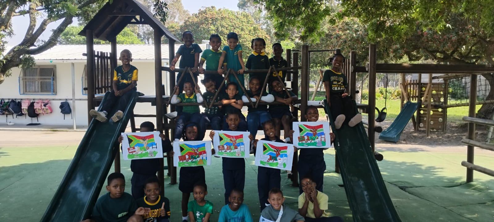 A group of children pose on and around a playground slide, some holding up colorful drawings. The setting is outdoors, with trees and a building in the background.