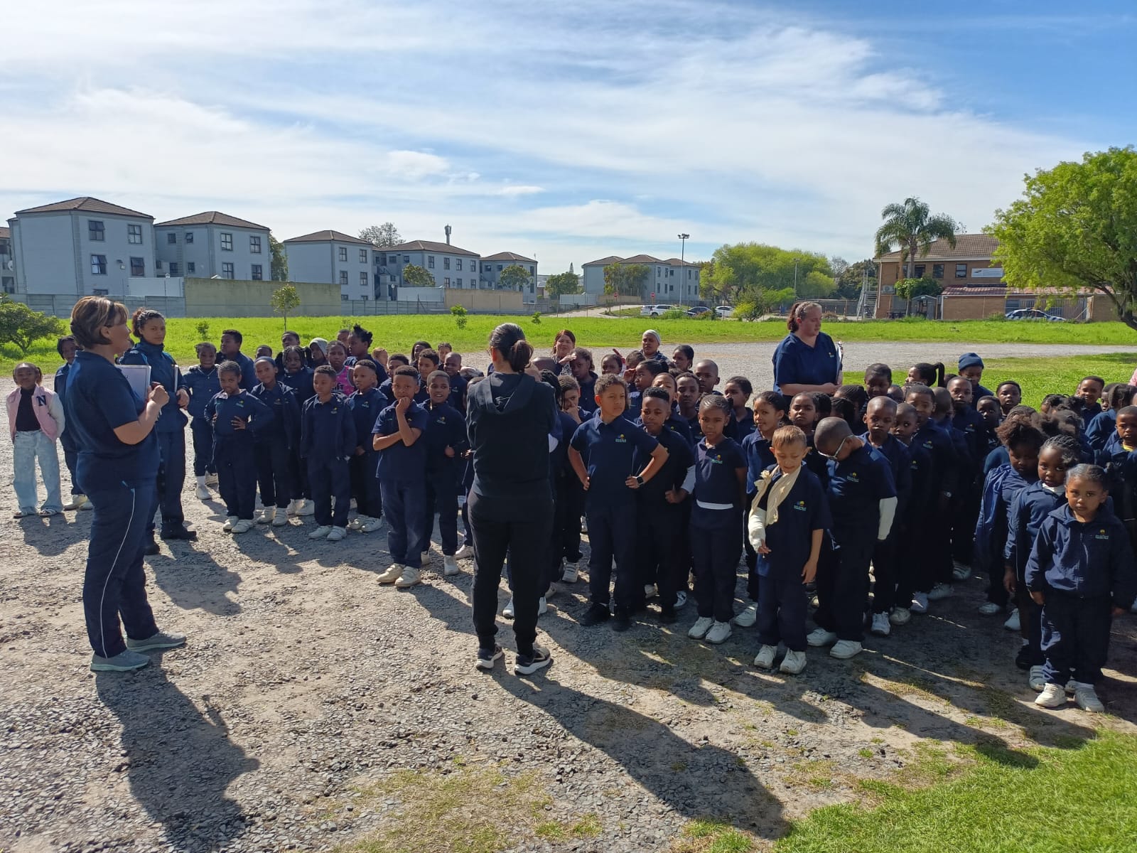 A group of children in navy uniforms stand together outdoors on a sunny day, with several adults nearby. Apartment buildings and trees are visible in the background.