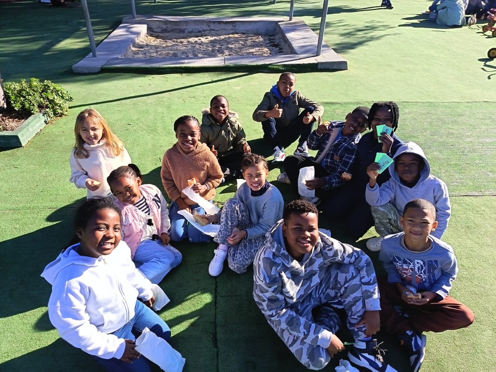 A group of children sit in a circle on a green playground surface, smiling and holding paper bags. There is a sandbox in the background and sunlight creates shadows on the ground.