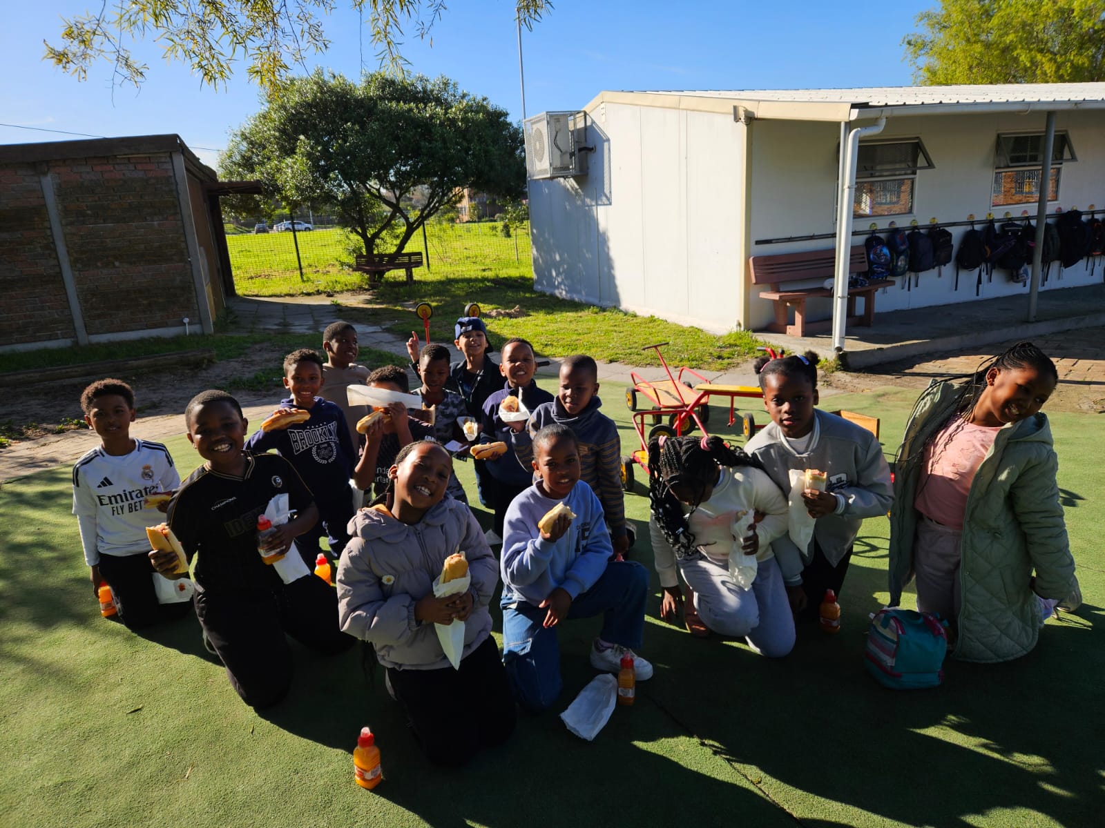 A group of children kneels outdoors on artificial grass, posing for a photo while holding sandwiches and juice bottles. School bags hang on hooks outside a nearby building. It is sunny with trees in the background.
