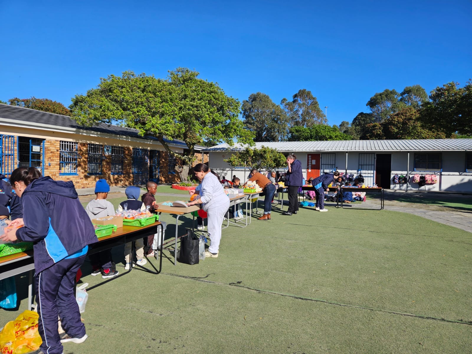 People stand and sit at tables outdoors on a sunny day, working with food items or produce. The area is covered in artificial grass, with buildings, trees, and blue sky in the background.