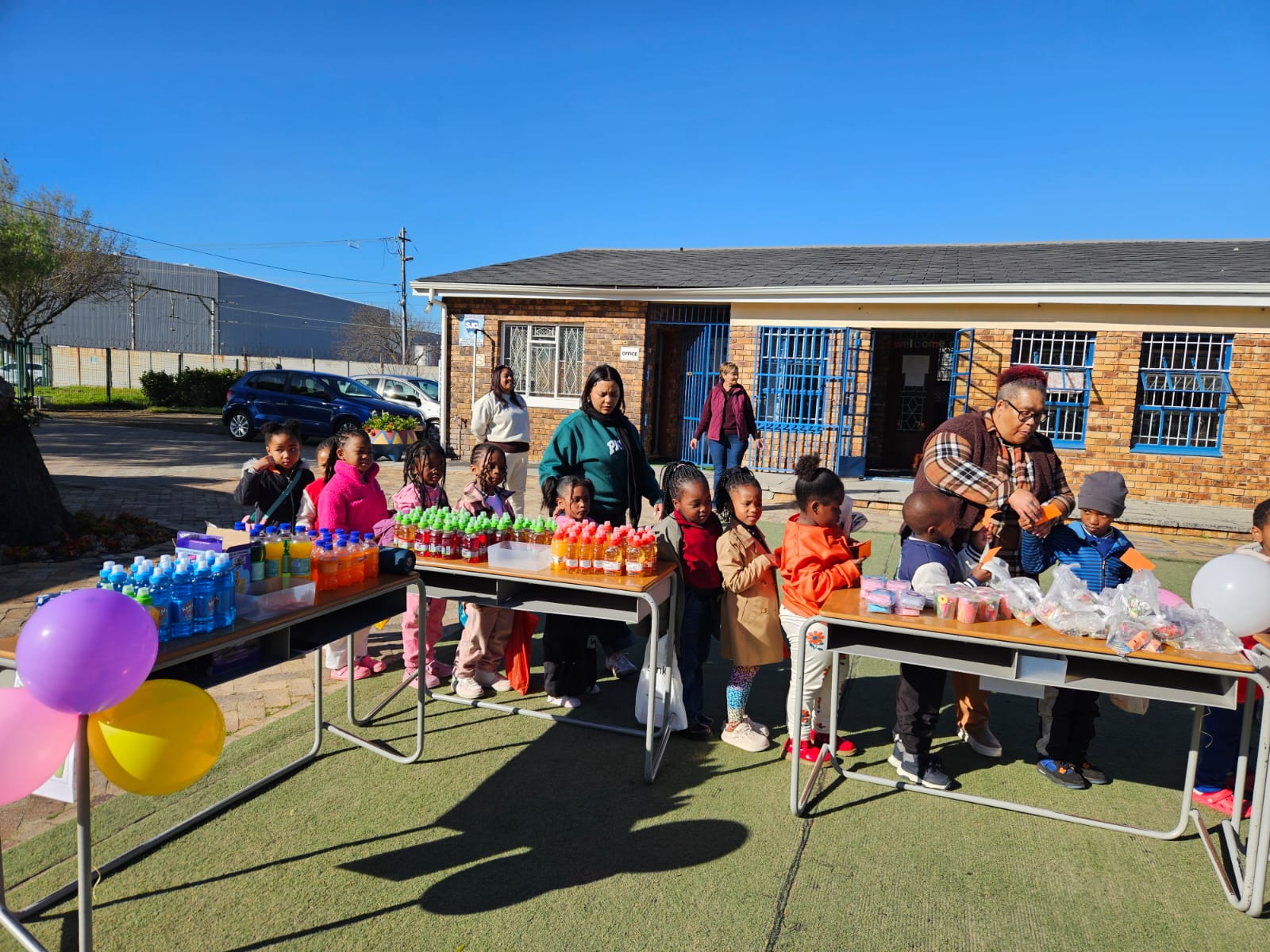 Children and adults stand behind tables with colorful drinks, snacks, and balloons outside a brick building on a sunny day. The group appears to be at an outdoor event or celebration.