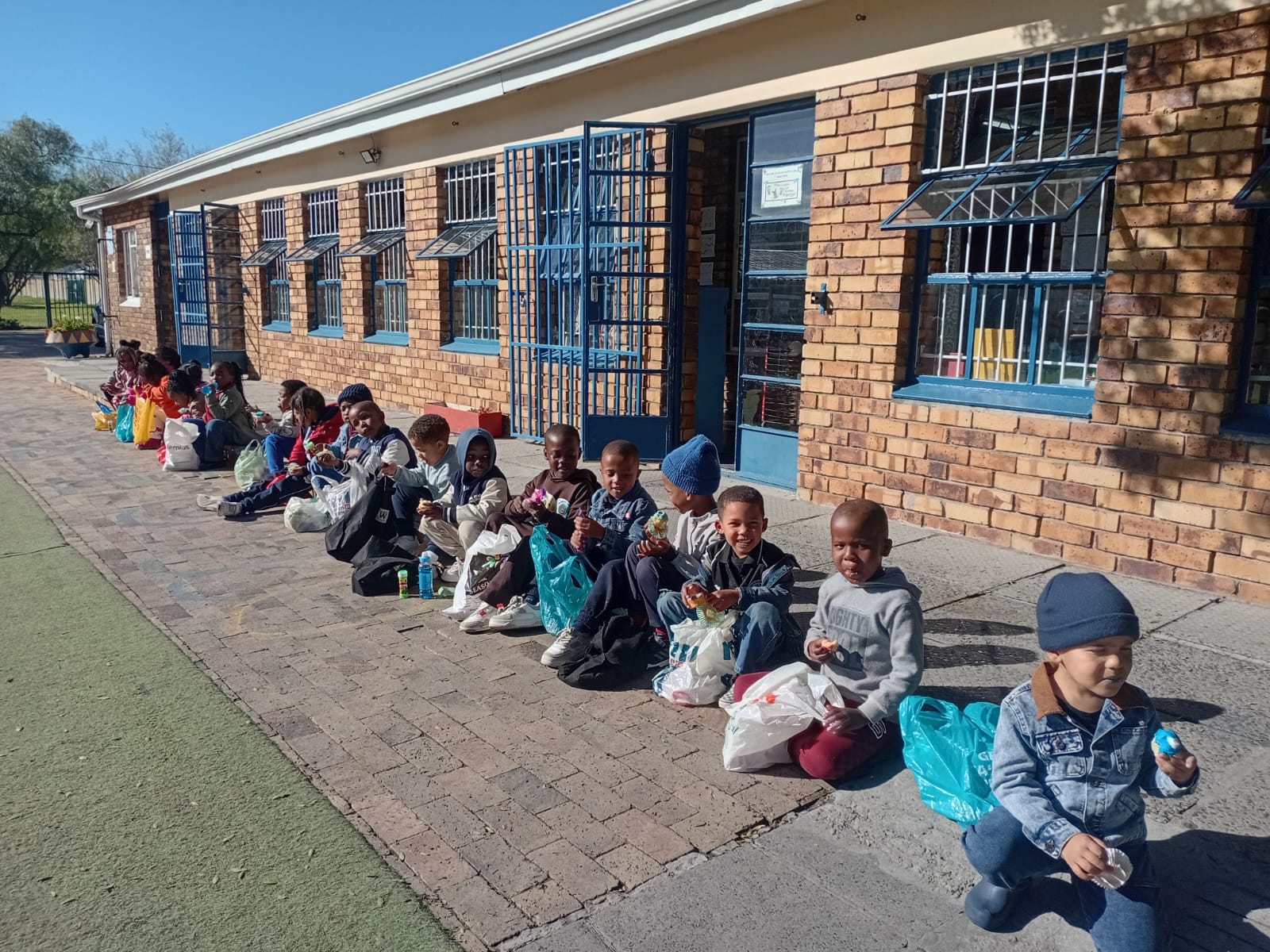 A group of children sit in a row outside a brick building, eating and holding drink bottles. Most have bags next to them. The weather appears sunny and clear. The building has blue doors and windows with bars.