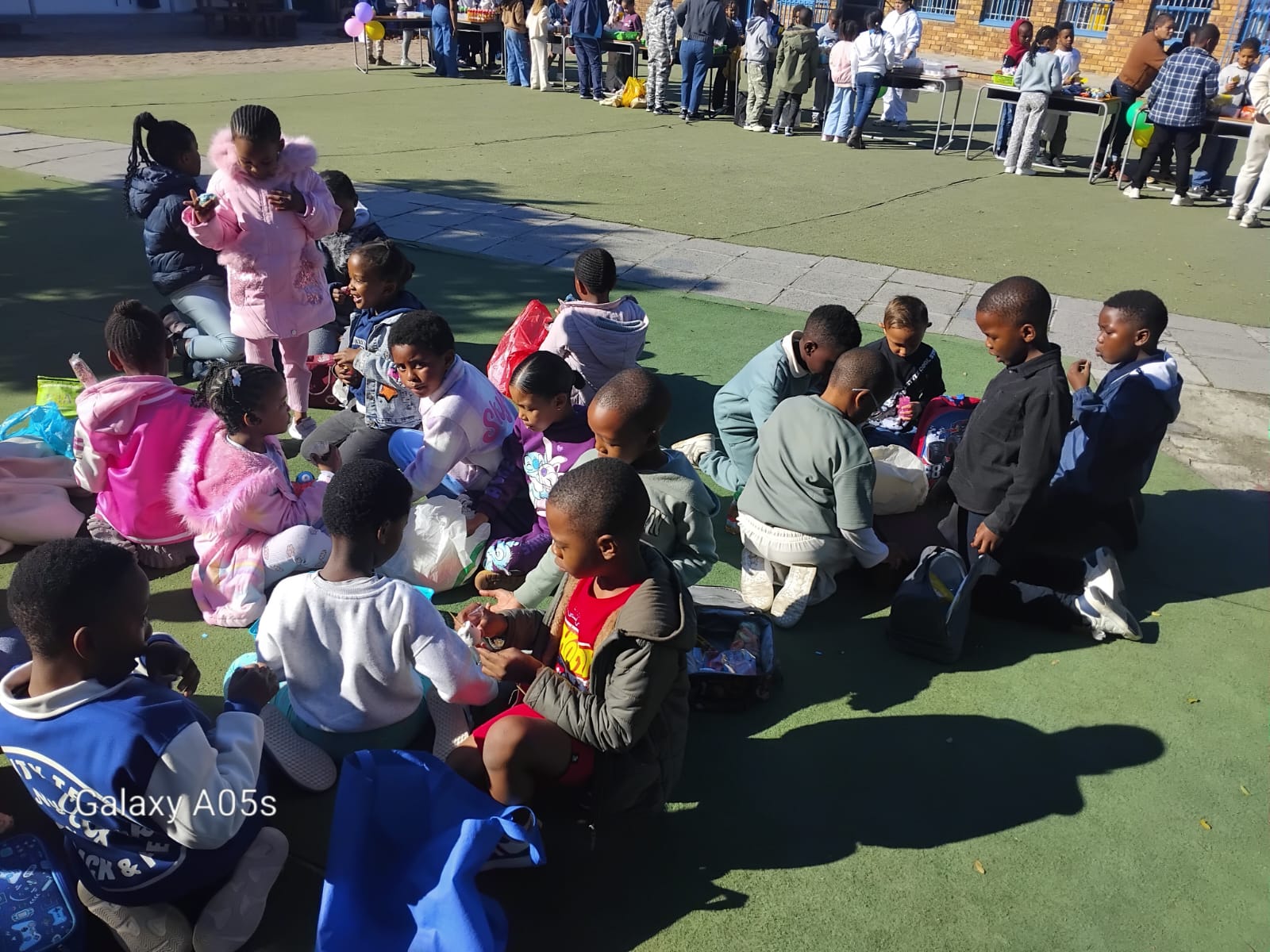 A group of children sit in circles on a green surface outdoors, eating and talking. Some adults stand nearby. More people are gathered in the background near tables. The scene is bright and sunny.