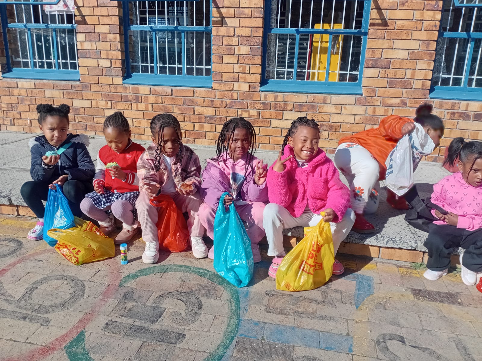 A group of young children sit on a pavement outside a brick building, holding colorful plastic bags and some snacks. They are dressed in bright clothes and appear to be posing for a photo.